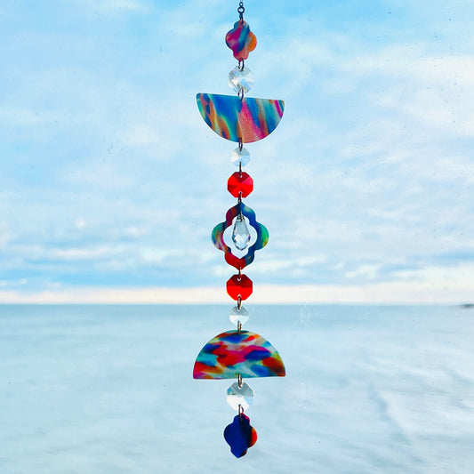 A handmade polymer clay suncatcher featuring marbled rainbow clay pieces in half-circle and quatrefoil shapes, separated by faceted clear glass beads and colorful accents. The suncatcher hangs vertically from a gold chain and includes a central drop-shaped crystal and red 14mm octagon crystals. It is photographed in front of a bright blue sky and calm lake horizon, with sunlight highlighting the bright swirls of red, blue, orange, yellow, and green in the clay.  