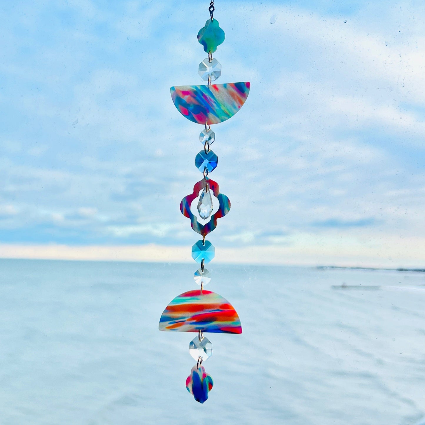 A handmade polymer clay suncatcher featuring marbled rainbow clay pieces in half-circle and quatrefoil shapes, separated by faceted clear glass beads and colorful accents. The suncatcher hangs vertically from a gold chain and includes a central drop-shaped crystal with blue & aqua 14mm octagon crystals. It is photographed in front of a bright blue sky and calm lake horizon, with sunlight highlighting the bright swirls of red, blue, orange, yellow, and green in the clay.